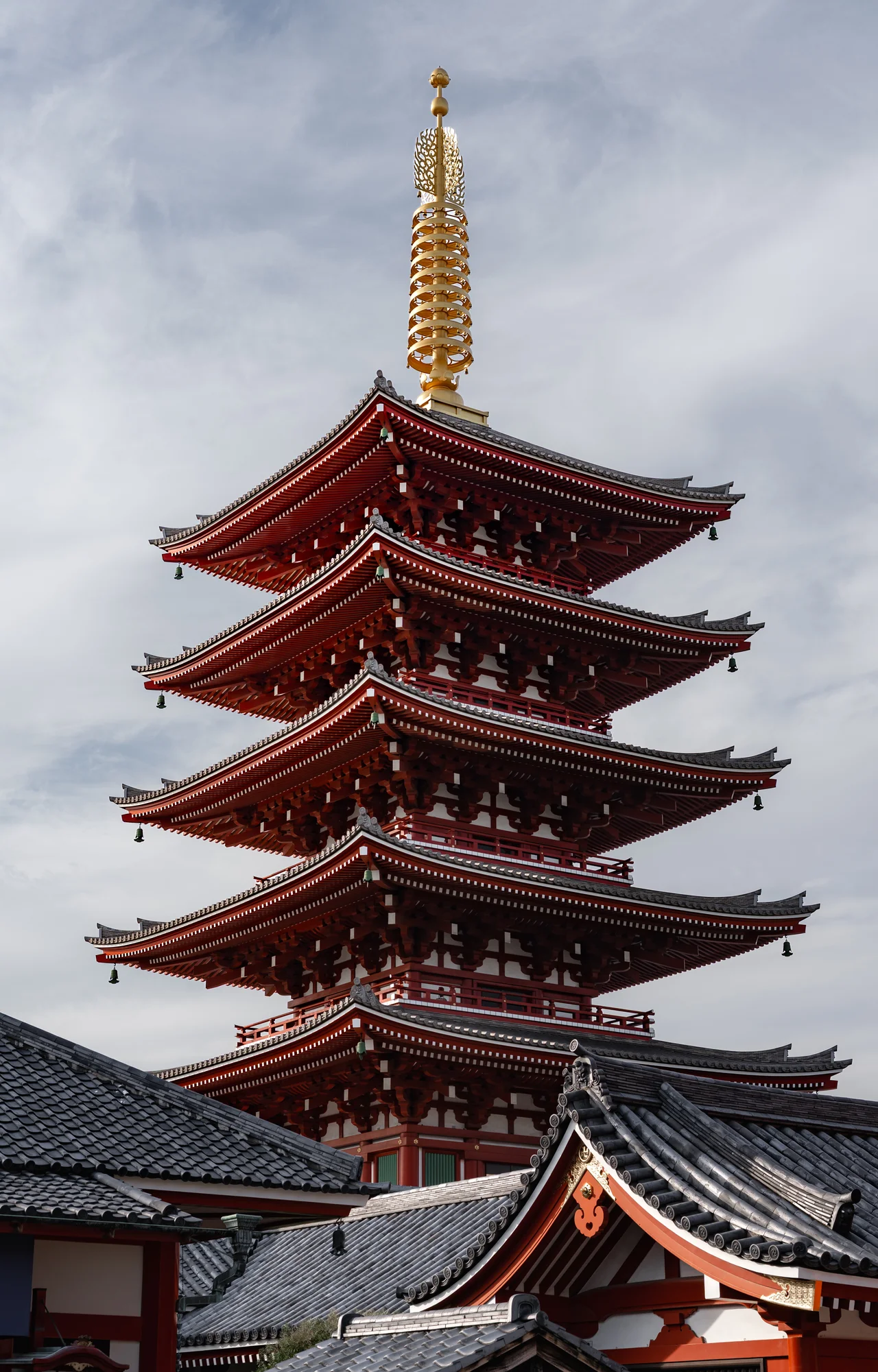 Pagode rouge à cinq étages surmontée d'une flèche dorée, au-dessus des toits d'un temple, sous un ciel nuageux.